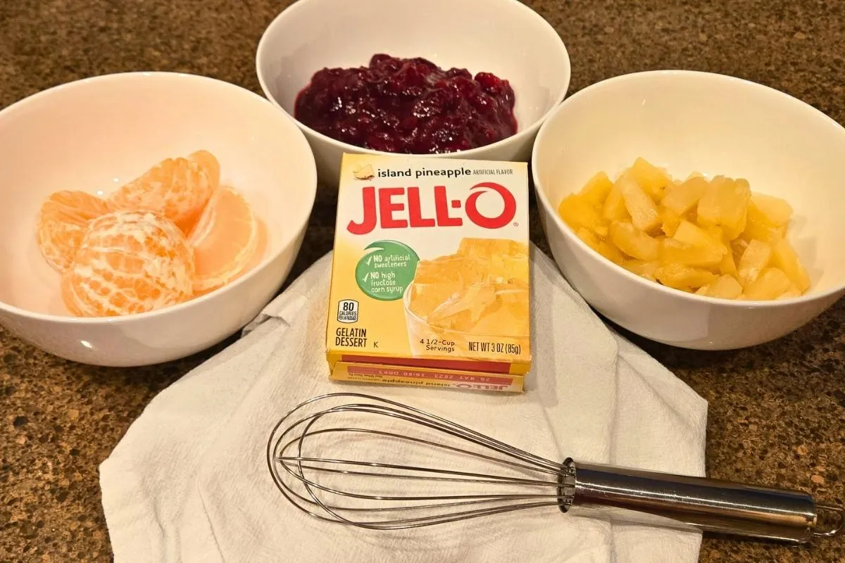 Prepared ingredients for a Jell-O dessert, including sliced mandarin oranges, diced pineapple, cranberry sauce, a box of Jell-O island pineapple flavor gelatin, and a whisk.