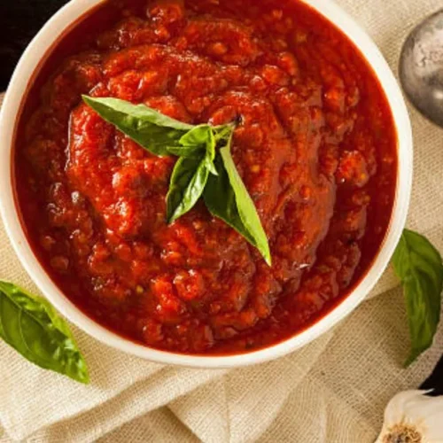 Homemade marinara sauce in a white bowl, garnished with fresh basil leaves, with whole tomatoes and garlic cloves in the background.