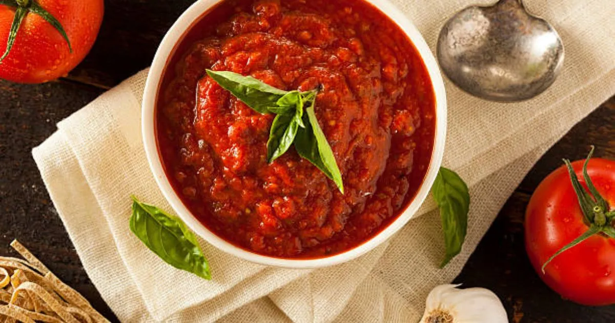 Homemade marinara sauce in a white bowl, garnished with fresh basil leaves, with whole tomatoes and garlic cloves in the background.