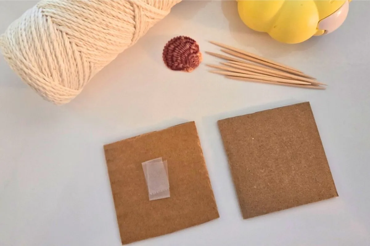 Craft supplies including a roll of twine, a seashell, toothpicks, and two pieces of cardboard with adhesive tape.
