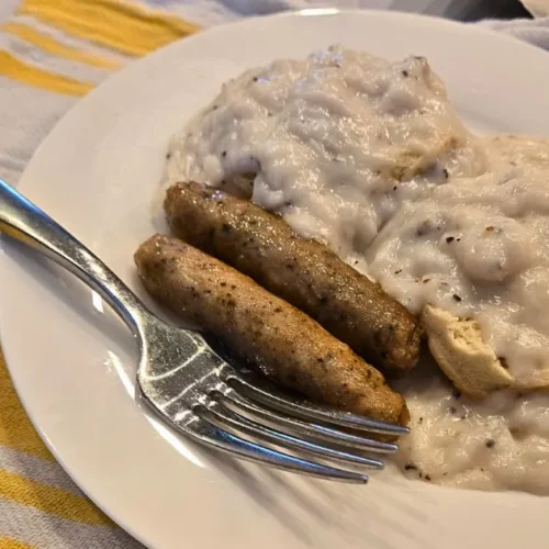 Biscuits smothered in sawmill gravy, alongside two breakfast sausages, on a white plate.