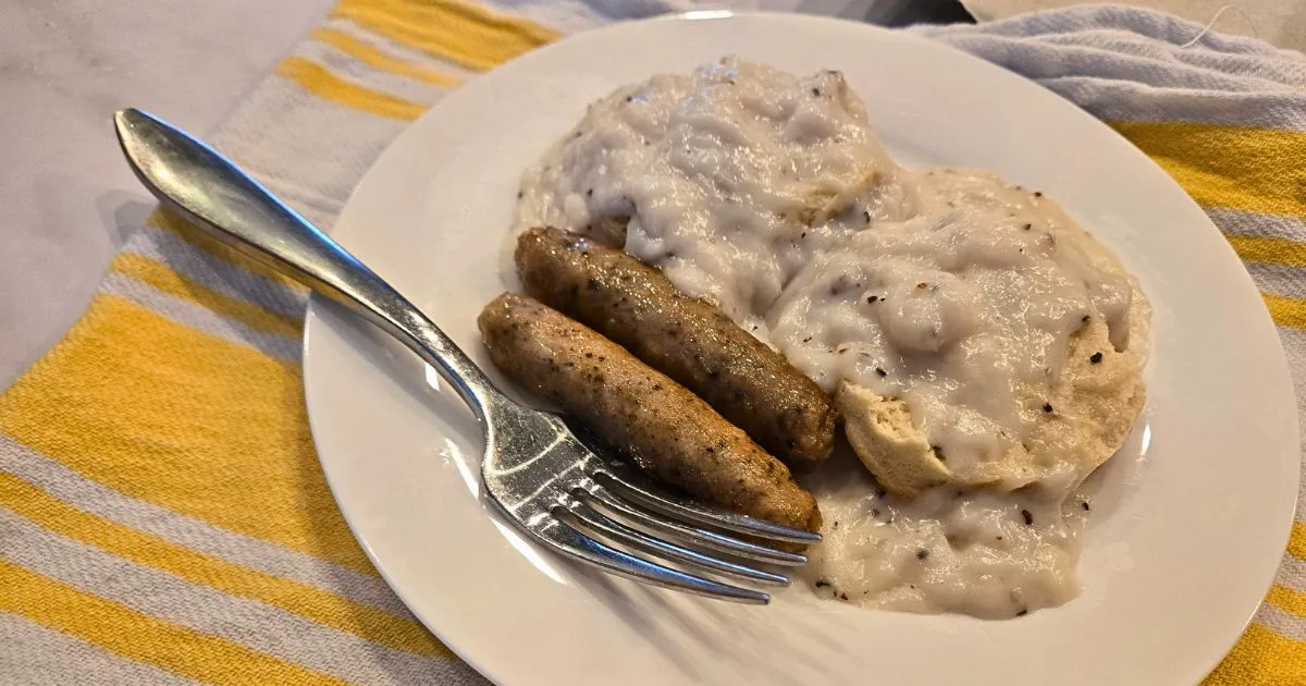 Biscuits smothered in sawmill gravy, alongside two breakfast sausages, on a white plate.