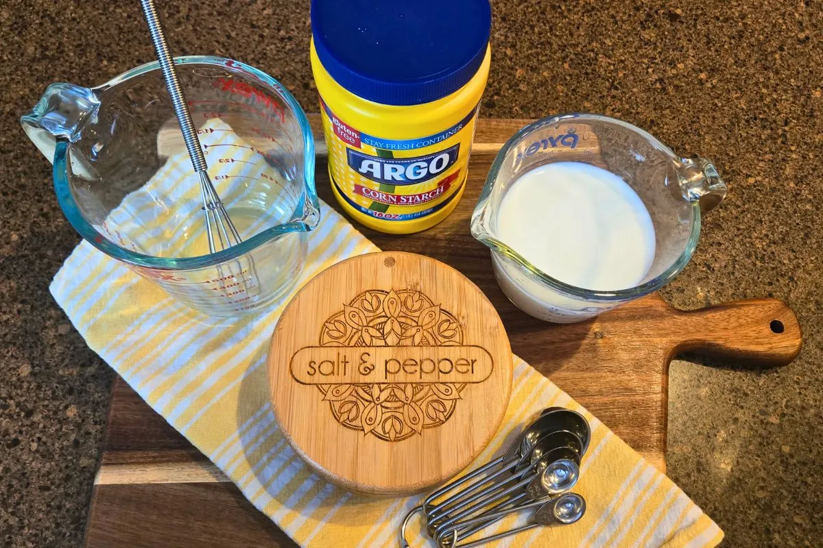 Measuring cups and spoons, corn starch, and milk, arranged on a wooden cutting board, ready for a microwave sawmill gravy recipe.