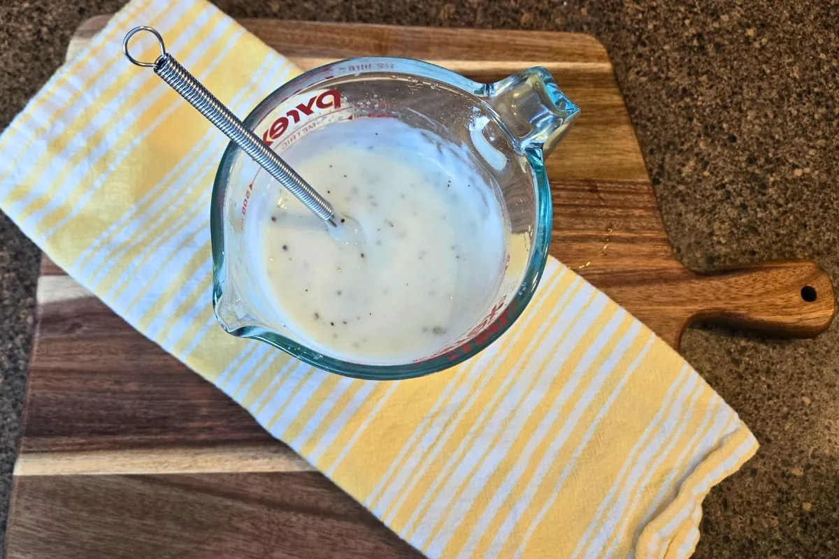 Measuring cup of microwave sawmill gravy with a whisk, on a wooden cutting board and yellow striped dishtowel.