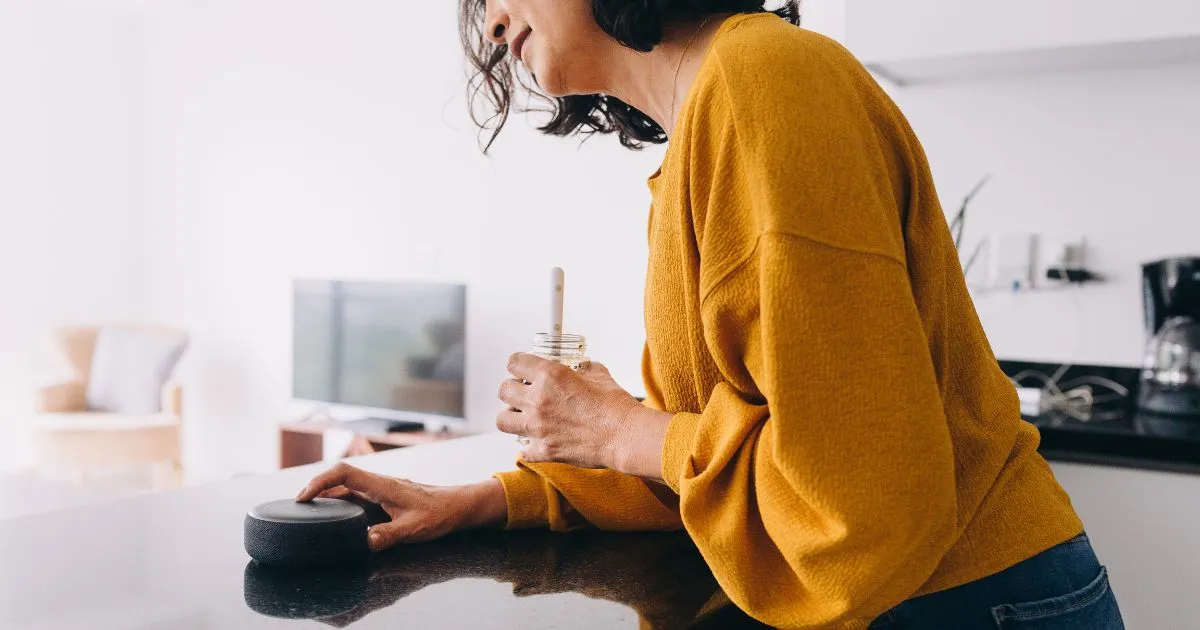 Woman in a mustard yellow sweater setting up an Alexa routine in a kitchen.
