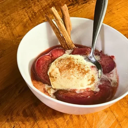 Baked plums served warm with vanilla ice cream and cinnamon sticks in a white bowl on a wooden table.