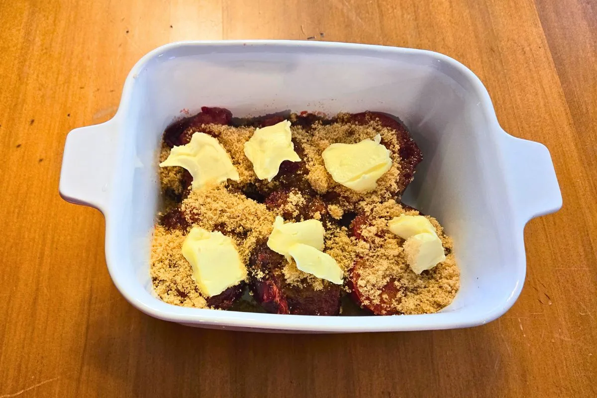 Halved red plums in a white baking dish topped with brown sugar and butter, ready for baking.