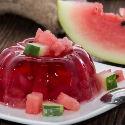 Red gelatin fruit salad topped with diced watermelon, served on a white plate with a spoon, and a slice of fresh watermelon in the background.