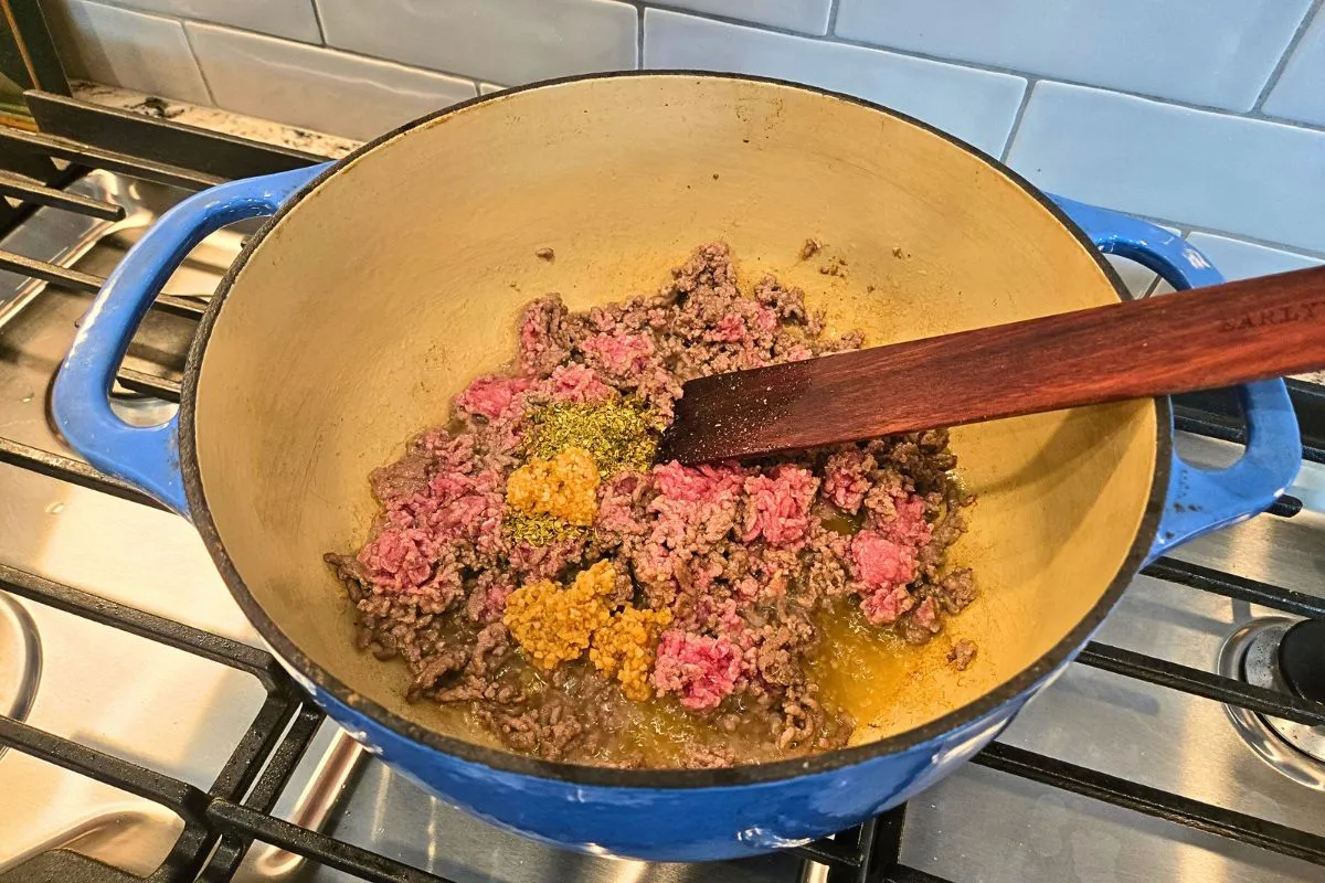 Ground beef browning in a blue Dutch oven with garlic and Italian seasoning for one pot lasagna