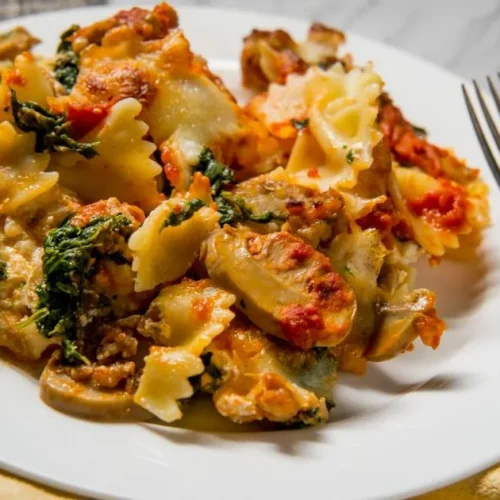 A serving of baked bowtie pasta with spinach, mushrooms, tomato sauce, and melted cheese on a white plate with a fork and spoon beside it.