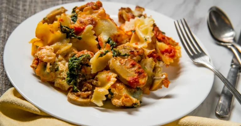 A serving of baked bowtie pasta with spinach, mushrooms, tomato sauce, and melted cheese on a white plate with a fork and spoon beside it.