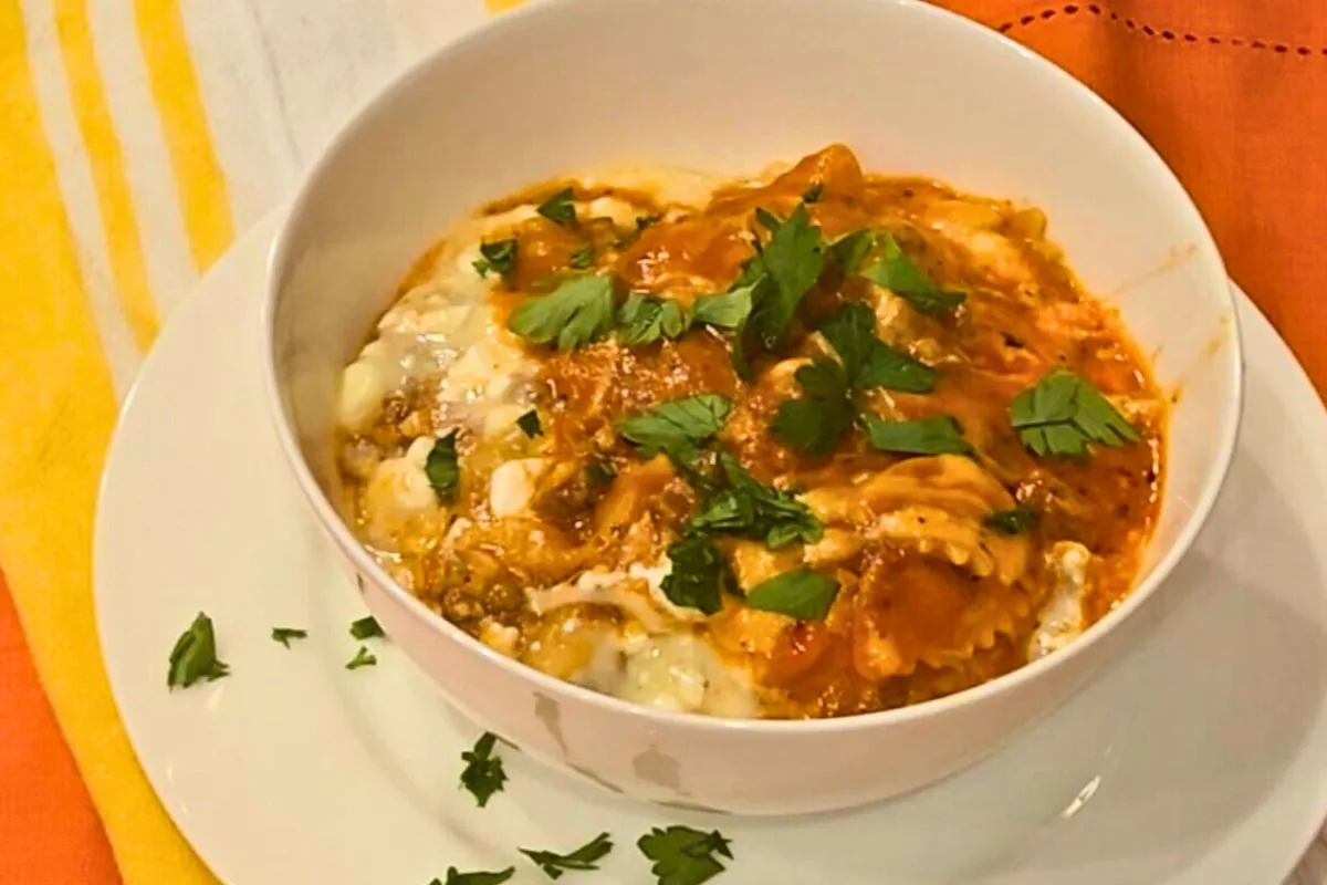 A white bowl of one-pot lasagna with pasta, melted cheese, and tomato sauce, garnished with fresh parsley, served on a white plate with an orange and yellow cloth in the background.