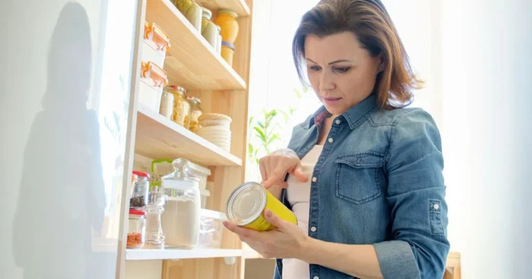 Woman standing in front of a pantry, wearing a denim shirt, holding a canned food item and reading the label. Shelves in the pantry are stocked with jars and containers of dry goods.