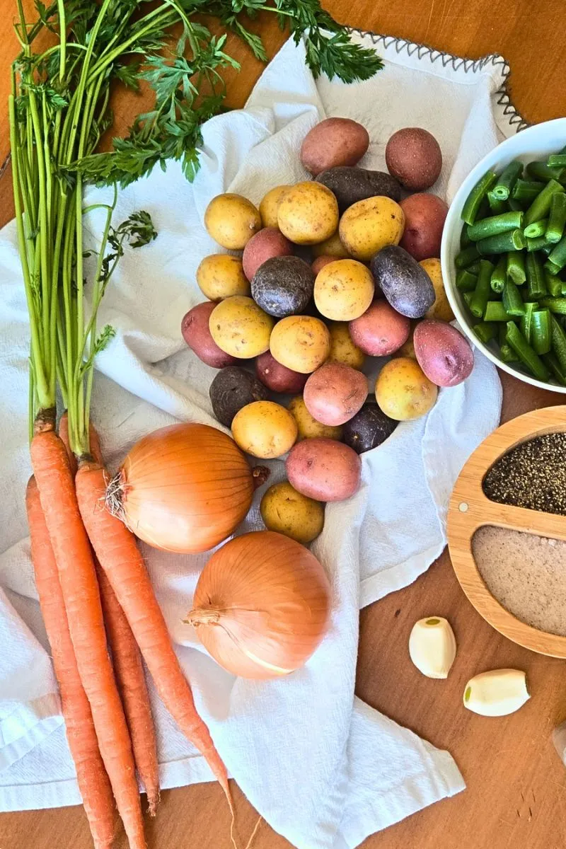 Overhead view of fresh carrots, onions, colorful baby potatoes, and green beans on a white cloth, arranged on a wooden kitchen table with garlic cloves and seasoning.