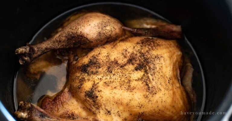A tender cooked whole chicken resting in a black slow-cooker style pot with juices in the bottom and seasoning on the skin.